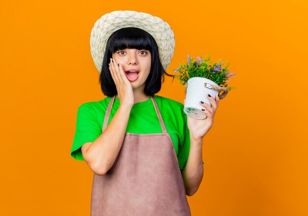 Jovem jardineira surpresa de uniforme, usando chapéu de jardinagem, segurando um vaso de flores