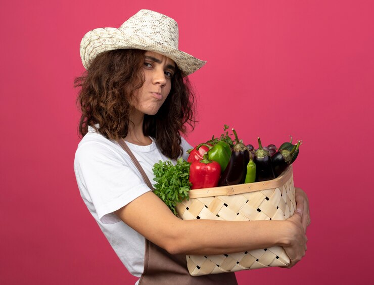 Foto grátis jovem jardineira triste de uniforme usando chapéu de jardinagem segurando uma cesta de vegetais isolada em rosa