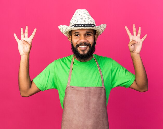 Jovem jardineiro afro-americano sorridente com chapéu de jardinagem mostrando quatro isolados na parede rosa