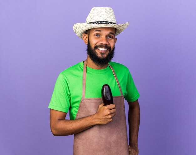 Jovem jardineiro afro-americano sorridente com chapéu de jardinagem segurando berinjela isolada na parede azul