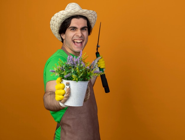 Foto grátis jovem jardineiro alegre de uniforme usando chapéu de jardinagem e luvas segurando uma tesoura e segurando uma flor em um vaso