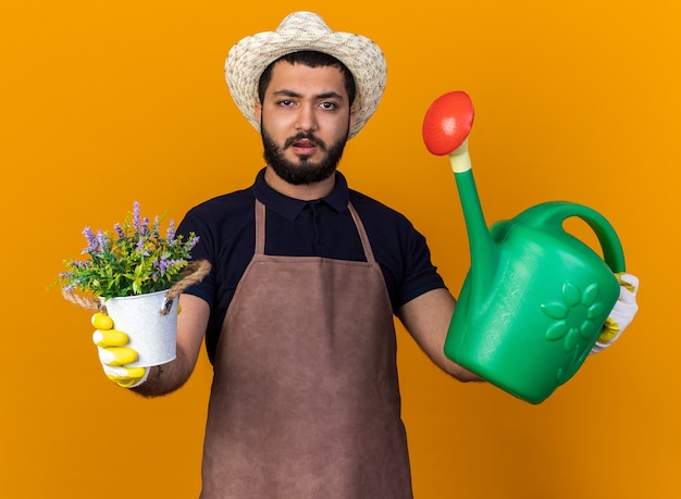 Jovem jardineiro caucasiano sem noção usando luvas e chapéu de jardinagem segurando um vaso de flores e um regador isolado em uma parede laranja com espaço de cópia