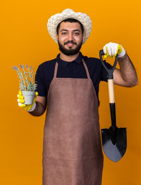 Jovem jardineiro caucasiano, sorridente, usando luvas e chapéu de jardinagem, segurando um vaso de flores e uma pá isolados na parede laranja com espaço de cópia