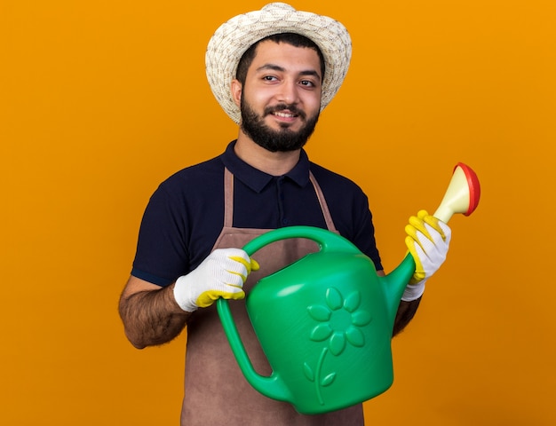 Jovem jardineiro caucasiano, sorridente, usando um chapéu de jardinagem e luvas segurando um regador, olhando para o lado isolado na parede laranja com espaço de cópia