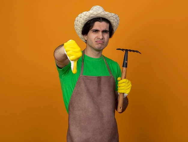 Foto grátis jovem jardineiro desagradável de uniforme, usando chapéu de jardinagem e luvas segurando um ancinho de enxada, mostrando o polegar isolado em laranja