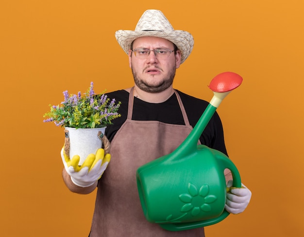 Jovem jardineiro descontente usando um chapéu de jardinagem e luvas segurando um regador e segurando uma flor em um vaso isolado na parede laranja