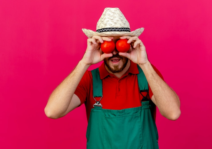 Foto grátis jovem jardineiro eslavo bonito de uniforme e chapéu segurando tomates na frente dos olhos