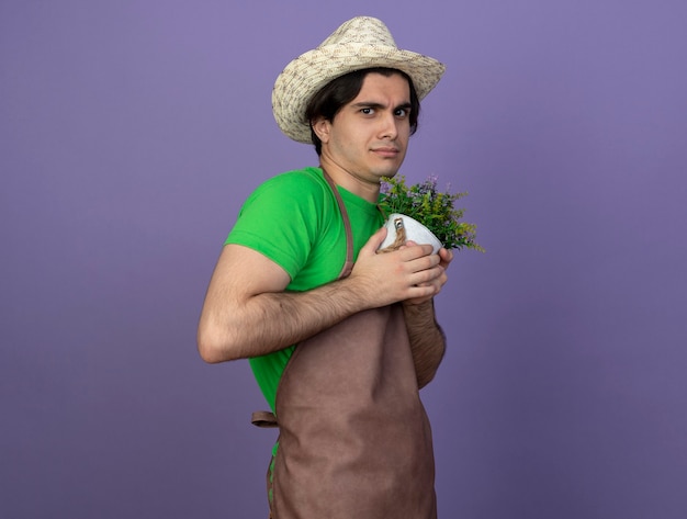 Jovem jardineiro ganancioso de uniforme, usando um chapéu de jardinagem, segurando uma flor em um vaso de flores