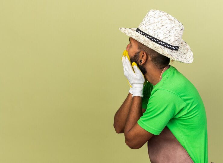 Foto grátis jovem jardineiro preocupado, cara afro-americana, usando chapéu de jardinagem com luvas, olhando para trás, isolado em uma parede verde oliva com espaço de cópia