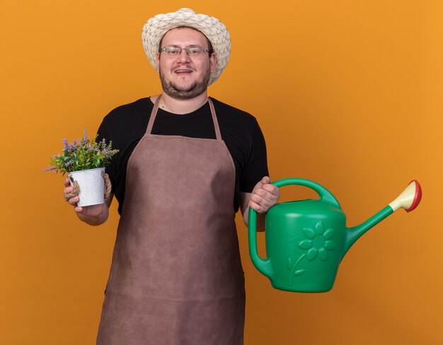 Jovem jardineiro satisfeito usando um chapéu de jardinagem e luvas, segurando um regador com uma flor em um vaso de flores isolado na parede laranja