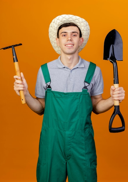 Jovem jardineiro sorridente com chapéu de jardinagem segura o ancinho e a pá isolados em um fundo laranja com espaço de cópia