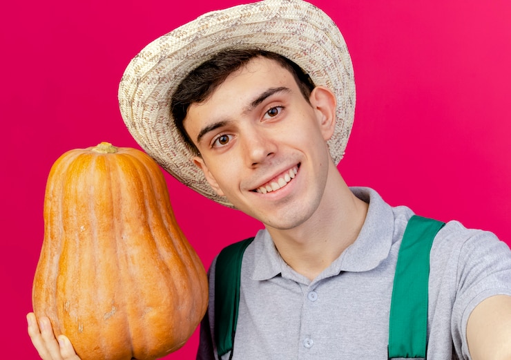 Foto grátis jovem jardineiro sorridente com chapéu de jardinagem segura uma abóbora e finge segurar uma câmera isolada em um fundo rosa com espaço de cópia