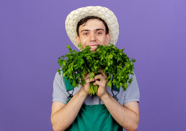 Jovem jardineiro sorridente com chapéu de jardinagem segurando coentro bem perto do rosto