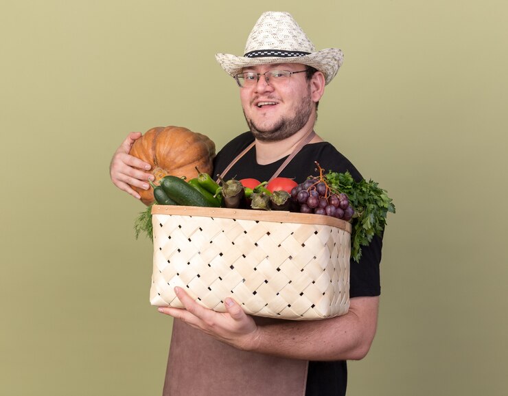 Foto grátis jovem jardineiro sorridente com chapéu de jardinagem segurando uma cesta de vegetais com abóbora isolada na parede verde oliva