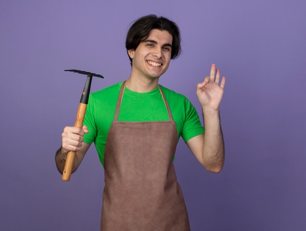 Foto grátis jovem jardineiro sorridente de uniforme segurando o ancinho e mostrando um gesto de aprovação