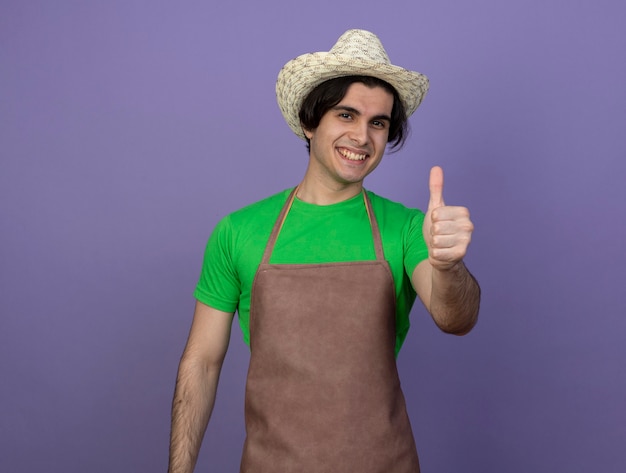 Jovem jardineiro sorridente de uniforme usando chapéu de jardinagem aparecendo o polegar