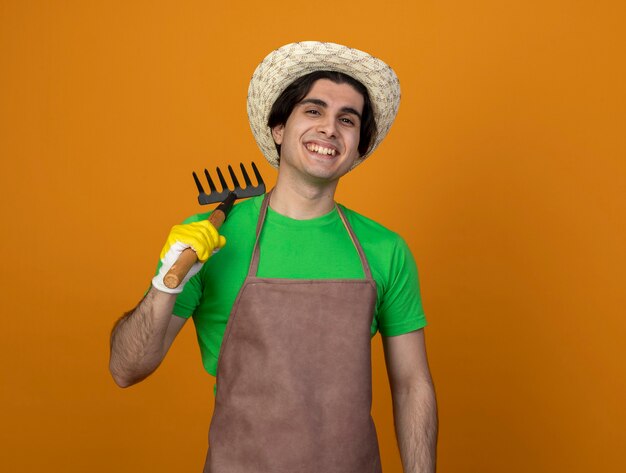 Jovem jardineiro sorridente de uniforme usando chapéu de jardinagem com luvas segurando o ancinho no ombro isolado em laranja
