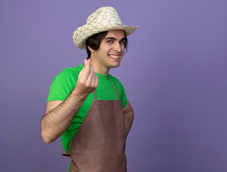 Foto grátis jovem jardineiro sorridente de uniforme usando chapéu de jardinagem mostrando gesto de gorjeta