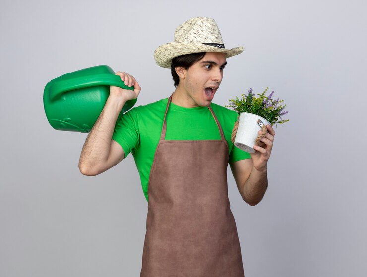Foto grátis jovem jardineiro surpreso de uniforme, usando chapéu de jardinagem, segurando um regador no ombro e olhando para uma flor em um vaso de flores