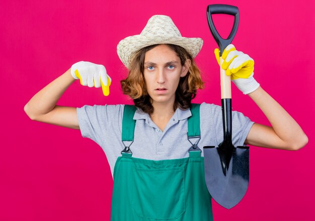 Foto grátis jovem jardineiro vestindo macacão e chapéu com luvas de borracha segurando uma pá