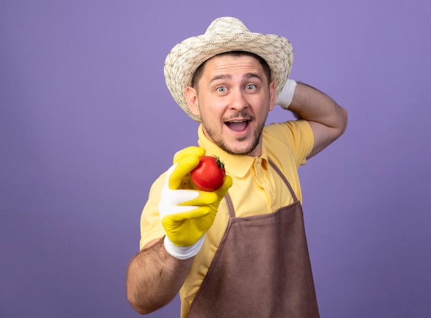 Jovem jardineiro vestindo macacão e chapéu em luvas de trabalho segurando tomate fresco feliz e positivo sorrindo alegremente