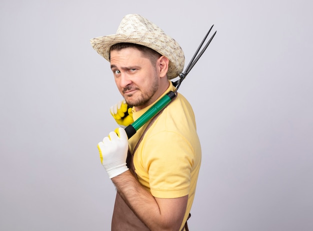 Jovem jardineiro vestindo macacão e chapéu em luvas de trabalho segurando um cortador de cerca viva olhando para a câmera sorrindo confiante