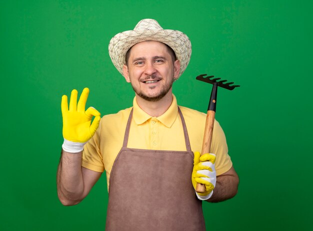 Jovem jardineiro vestindo macacão e chapéu em luvas de trabalho segurando um mini ancinho olhando para frente sorrindo mostrando uma placa de ok em pé sobre a parede verde