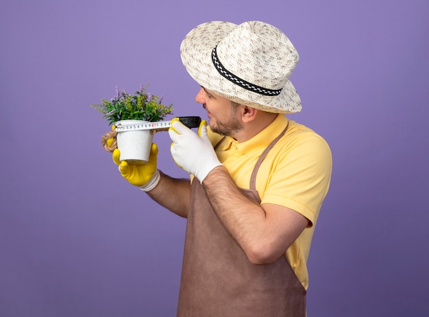 Jovem jardineiro vestindo macacão e chapéu em luvas de trabalho segurando um vaso de planta medindo-o com fita métrica, parecendo intrigado de pé sobre a parede roxa