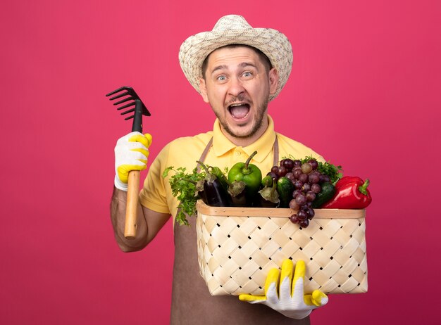 Jovem jardineiro vestindo macacão e chapéu em luvas de trabalho segurando uma caixa cheia de vegetais com um mini ancinho gritando animado e feliz em pé sobre a parede rosa