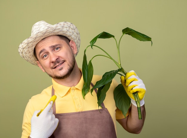 Jovem jardineiro vestindo macacão e chapéu em luvas de trabalho segurando uma planta olhando para a frente sorrindo com uma cara feliz em pé sobre a parede de luz