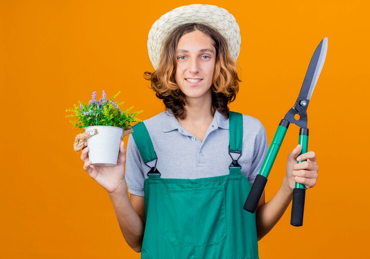 Foto grátis jovem jardineiro vestindo macacão e chapéu segurando um cortador de cerca viva e um vaso de plantas