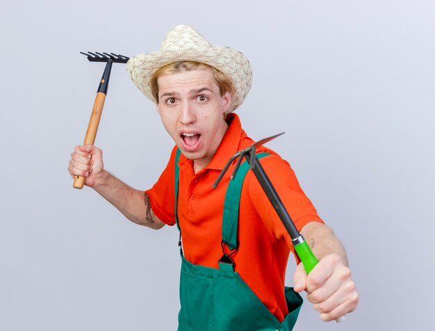 Jovem jardineiro vestindo macacão e chapéu segurando um mini ancinho e enxadão olhando para a câmera gritando com expressão de medo em pé sobre um fundo branco