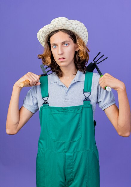 Jovem jardineiro vestindo macacão e chapéu segurando um mini ancinho e enxadão