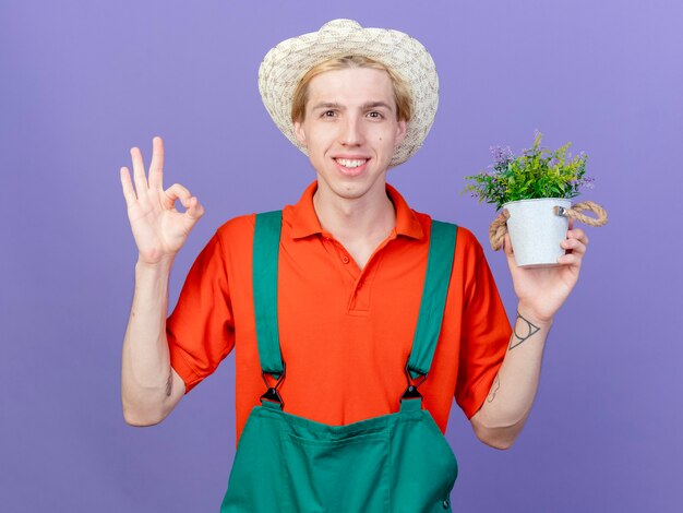 Jovem jardineiro vestindo macacão e chapéu segurando um vaso de plantas sorrindo