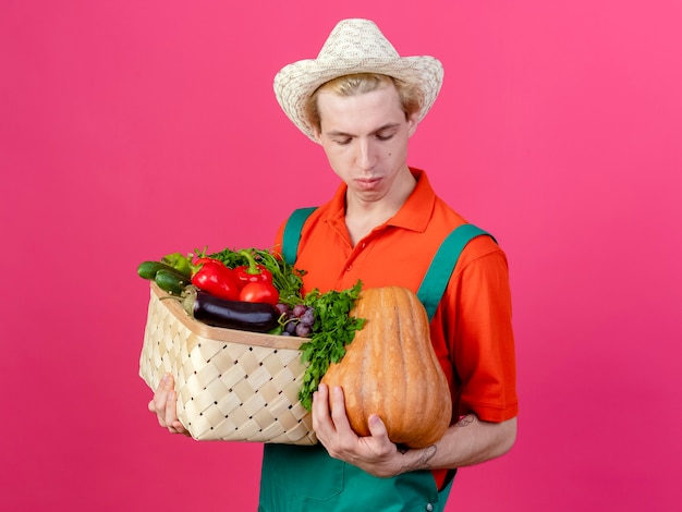 Jovem jardineiro vestindo macacão e chapéu segurando uma caixa cheia de vegetais