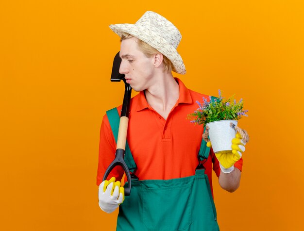 Jovem jardineiro vestindo macacão e chapéu segurando uma pá e um vaso de plantas olhando para o lado