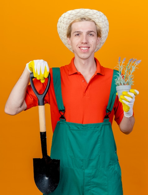 Jovem jardineiro vestindo macacão e chapéu segurando uma pá e um vaso de plantas