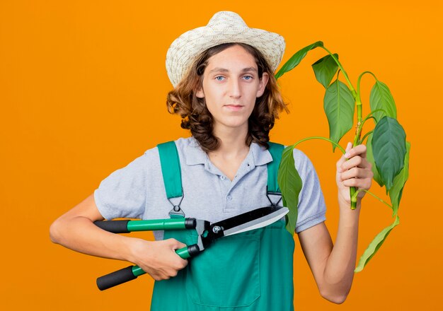Jovem jardineiro vestindo macacão e chapéu segurando uma planta e um cortador de cerca viva