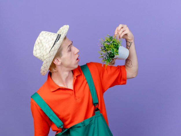 Jovem jardineiro vestindo macacão e chapéu segurando uma planta em um vaso olhando para ela surpresa