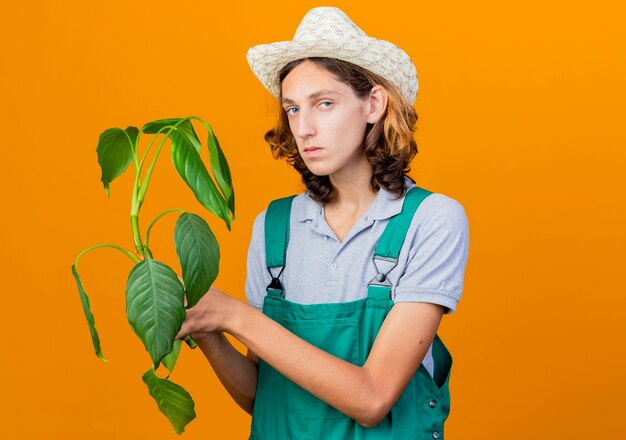 Jovem jardineiro vestindo macacão e chapéu segurando uma planta