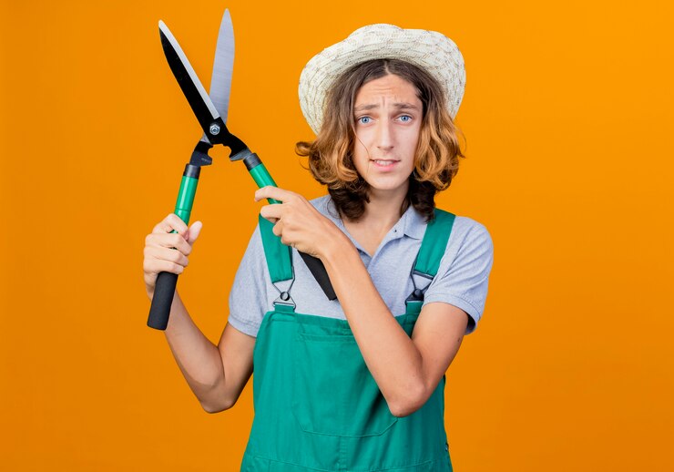 Foto grátis jovem jardineiro vestindo macacão e chapéu segurando uma tesoura de sebe sendo confuso