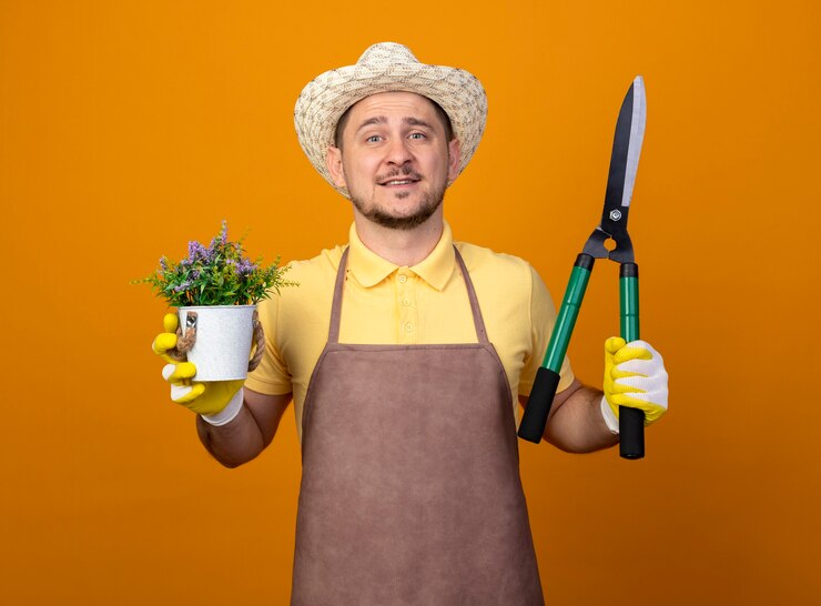 Foto grátis jovem jardineiro vestindo macacão e chapéu segurando uma tesoura de sebes e um vaso de plantas olhando para a frente com uma cara feliz sorrindo em pé sobre a parede laranja