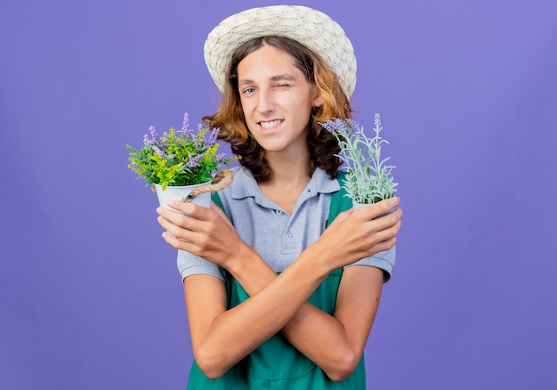Jovem jardineiro vestindo macacão e chapéu segurando vasos de plantas sorrindo