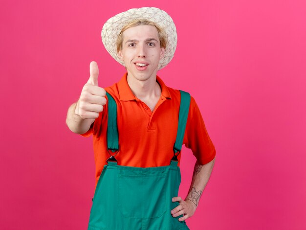 Jovem jardineiro vestindo macacão e chapéu sorrindo e mostrando os polegares para cima