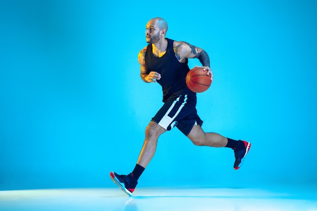 Jovem jogador de basquete da equipe vestindo treinamento sportwear, praticando em ação, movimento isolado sobre um fundo azul em luz de néon. conceito de esporte, movimento, energia e estilo de vida dinâmico e saudável.