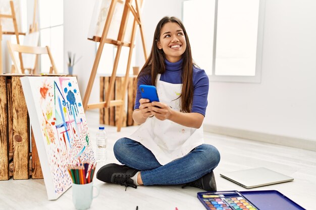 Jovem latina usando smartphone sentado no chão no estúdio de arte