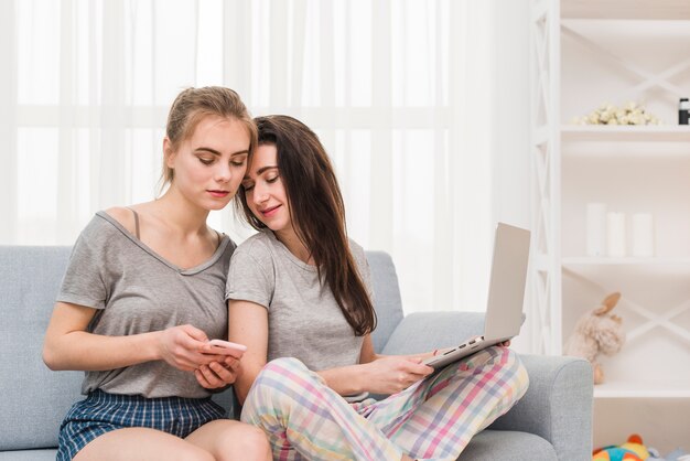 Jovem lésbica segurando laptop no colo olhando no celular da namorada