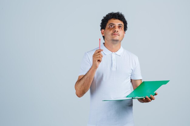 Jovem, levantando a caneta em gesto de eureka e segurando o caderno em jeans e camiseta branca e olhando pensativo. vista frontal.