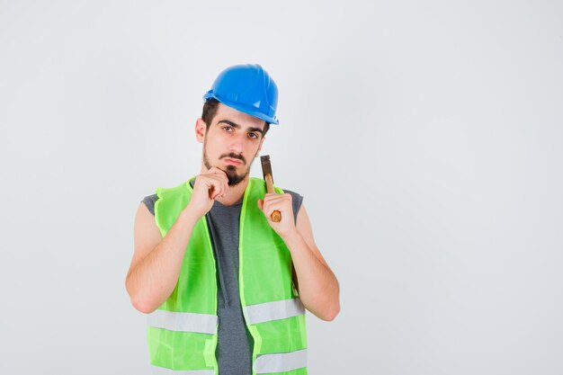 Jovem levantando o machado sobre o ombro e apoiando o queixo na mão em uniforme de construção e parecendo pensativo