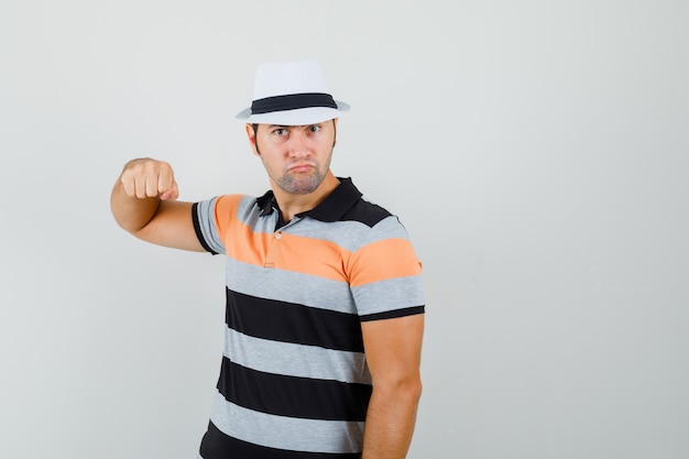 Foto grátis jovem levantando o punho em camiseta listrada, chapéu e olhando com raiva, vista frontal.